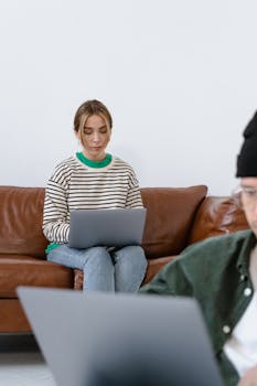Focused young woman using laptop on a brown sofa while working remotely indoors.