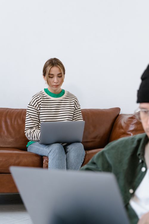 Free Focused young woman using laptop on a brown sofa while working remotely indoors. Stock Photo