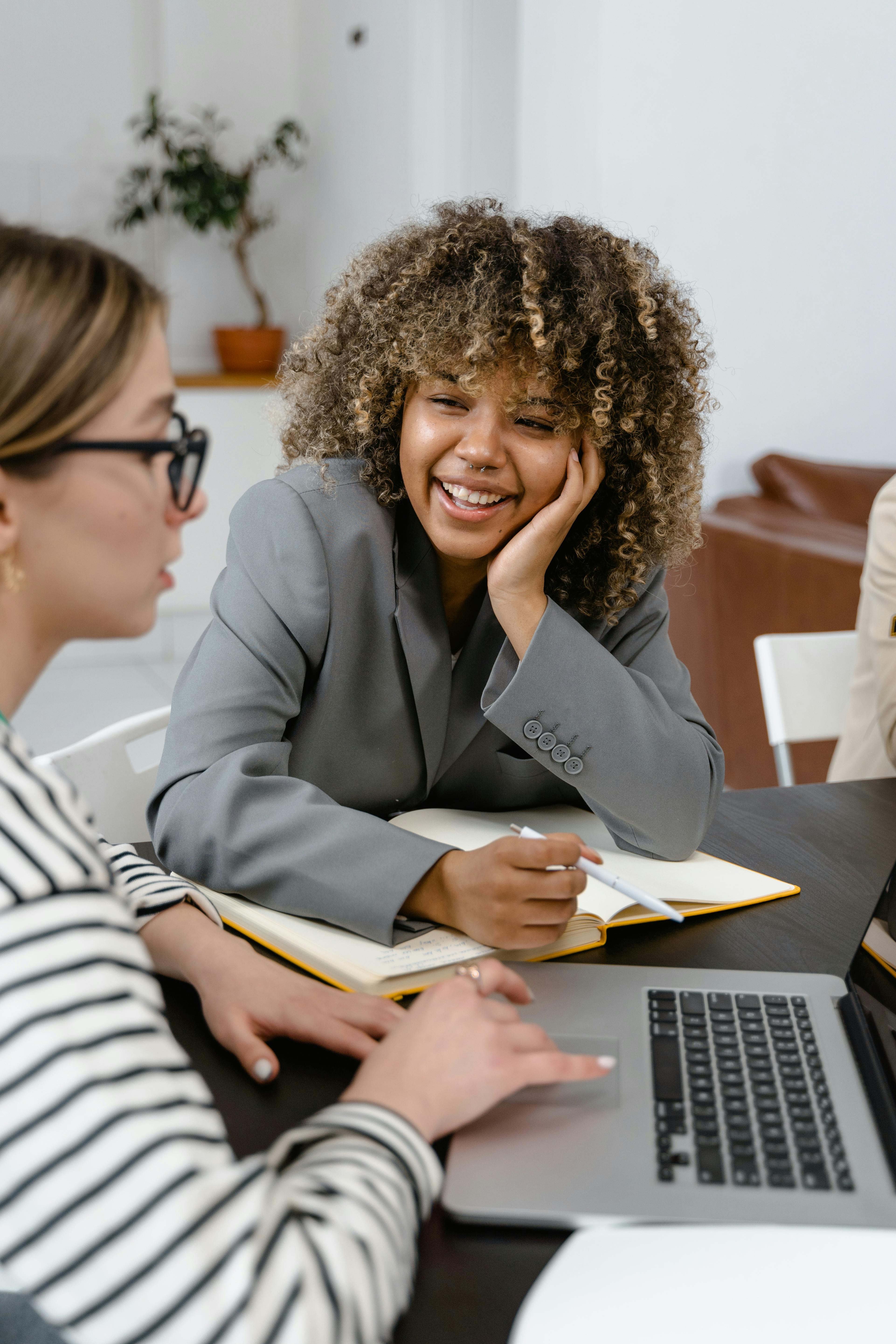 a woman in gray blazer smiling while looking at her colleague using a laptop