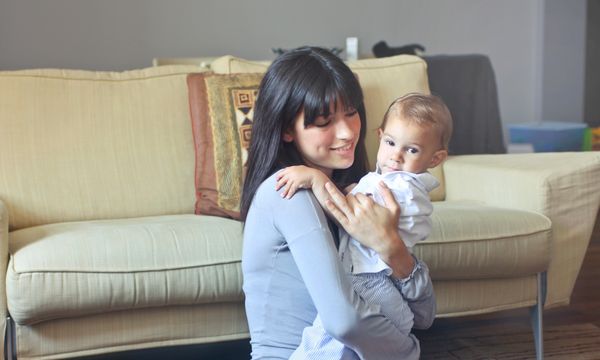 Nanny (Part-time) — A mother lovingly embraces her baby indoors on a comfortable sofa capturing warmth and togetherness.
