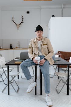 Man in casual attire sitting on a desk holding a cup in a stylish modern office with decor.