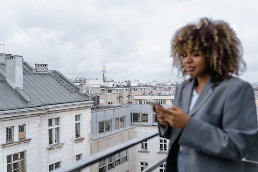 Blurry young woman with curly hair using a smartphone on a city balcony.