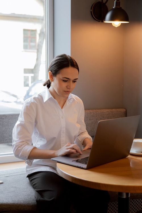 Free Caucasian woman in white blouse working on laptop at a cafe table during the day. Stock Photo