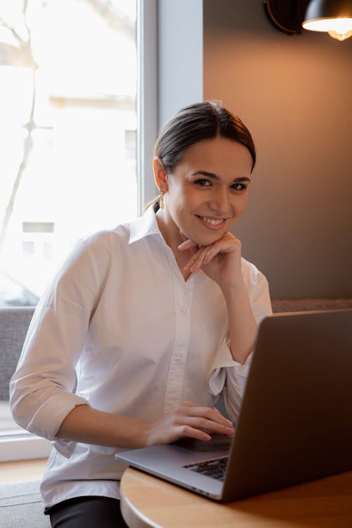 Free Woman in a White Dress Shirt Smiling Near Her Laptop Stock Photo