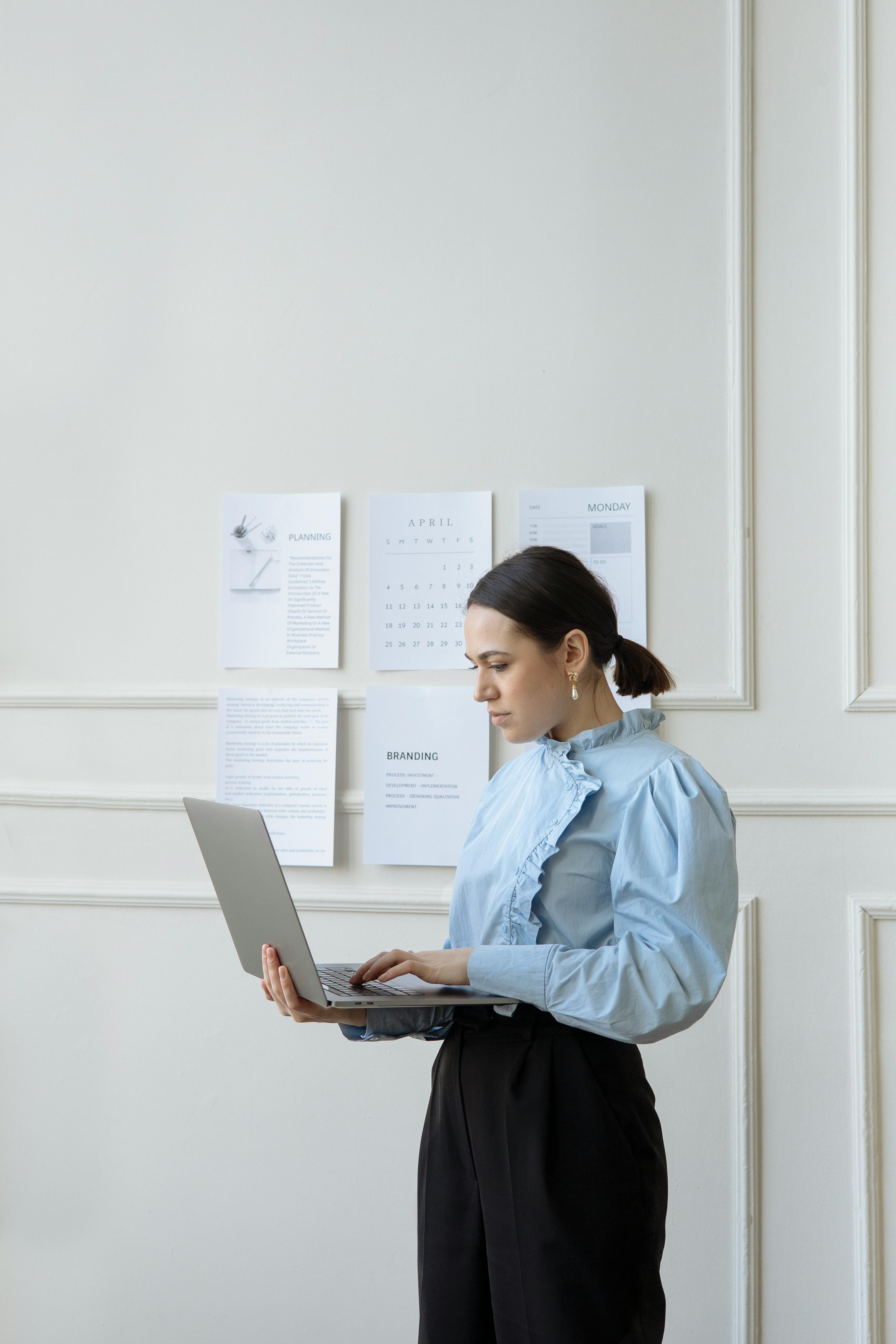 a woman in blue long sleeves and black pants standing while working on her laptop