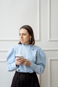 Young woman in blue shirt uses smartphone indoors, looking aside thoughtfully.