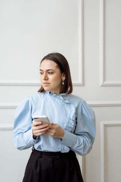 Free Young woman in blue shirt uses smartphone indoors, looking aside thoughtfully. Stock Photo