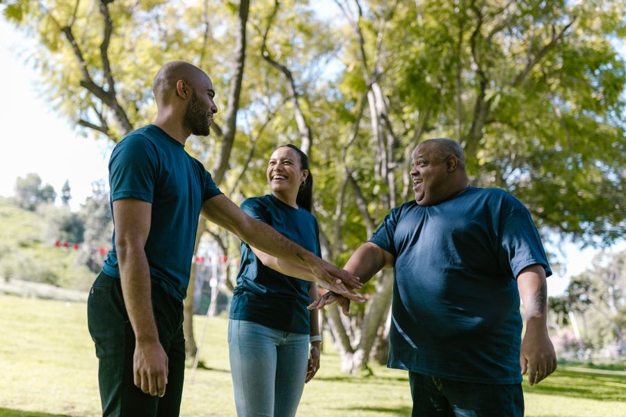 An adult with Down syndrome and two support professionals share a joyful moment outdoors, hands joined.