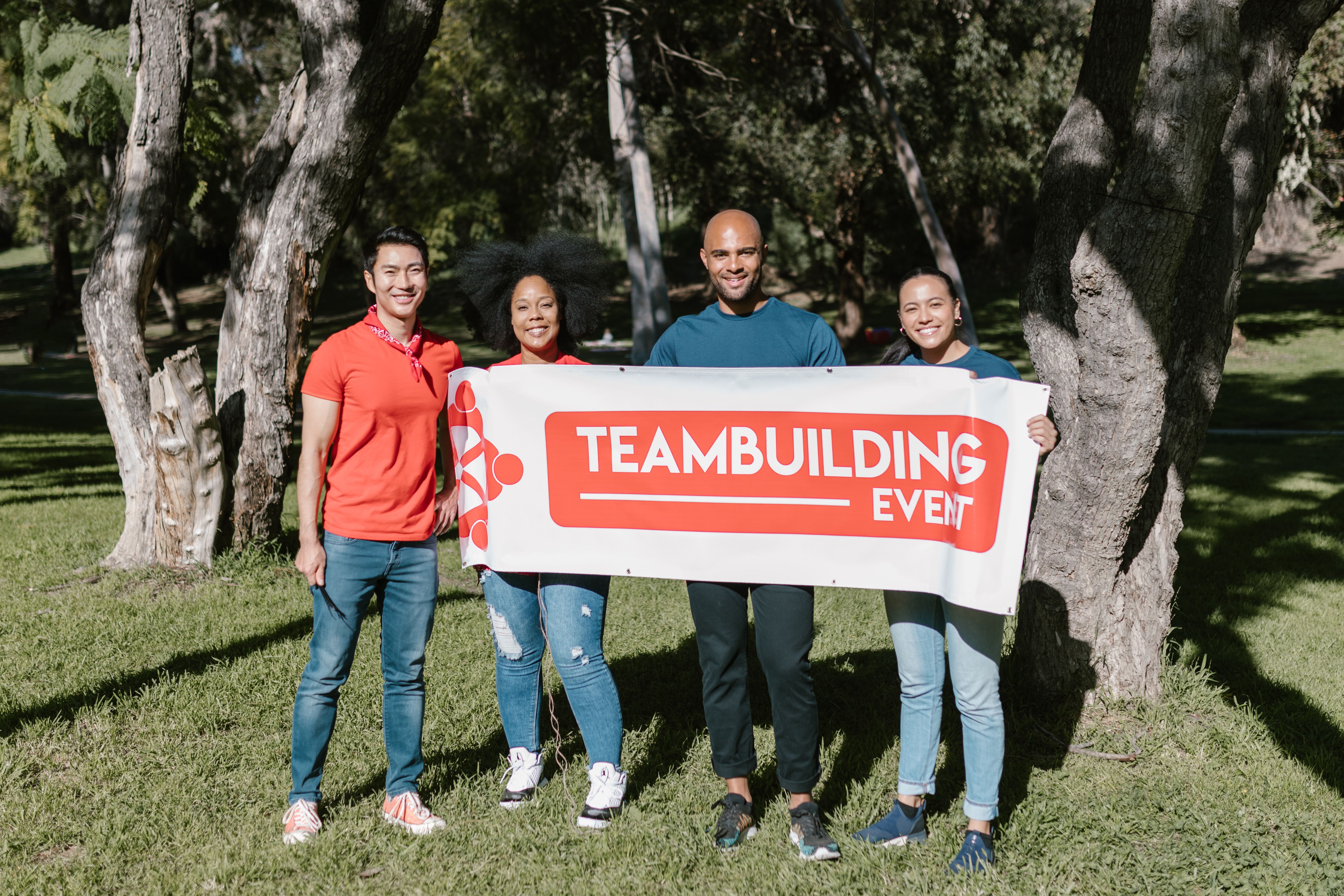 Group of four diverse adults holding a team building banner in a sunny park.
