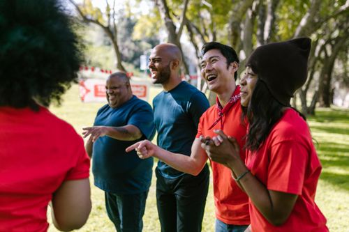 Un groupe diversifié de bénévoles, vêtus de t-shirts rouges, rit et discute joyeusement dans un parc arboré en journée.