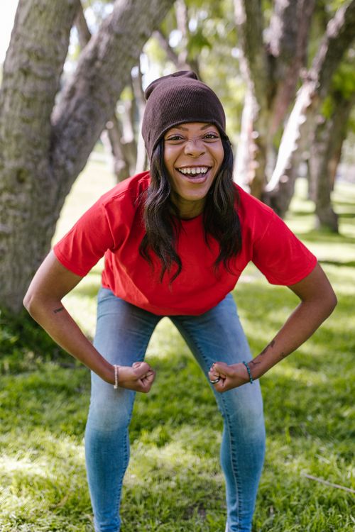 Free Young woman in a red shirt smiling energetically in a park, flexing her muscles. Stock Photo