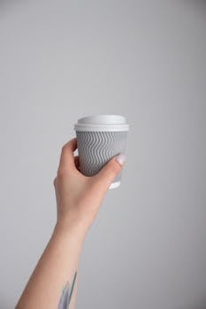 Minimalist photo of a hand holding a gray disposable coffee cup with a tattoo against a neutral background.