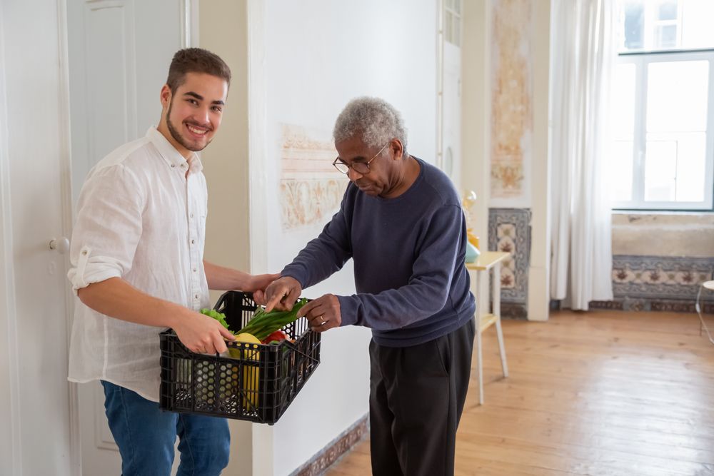 A helper bringing a crate of fresh groceries to an older man at home