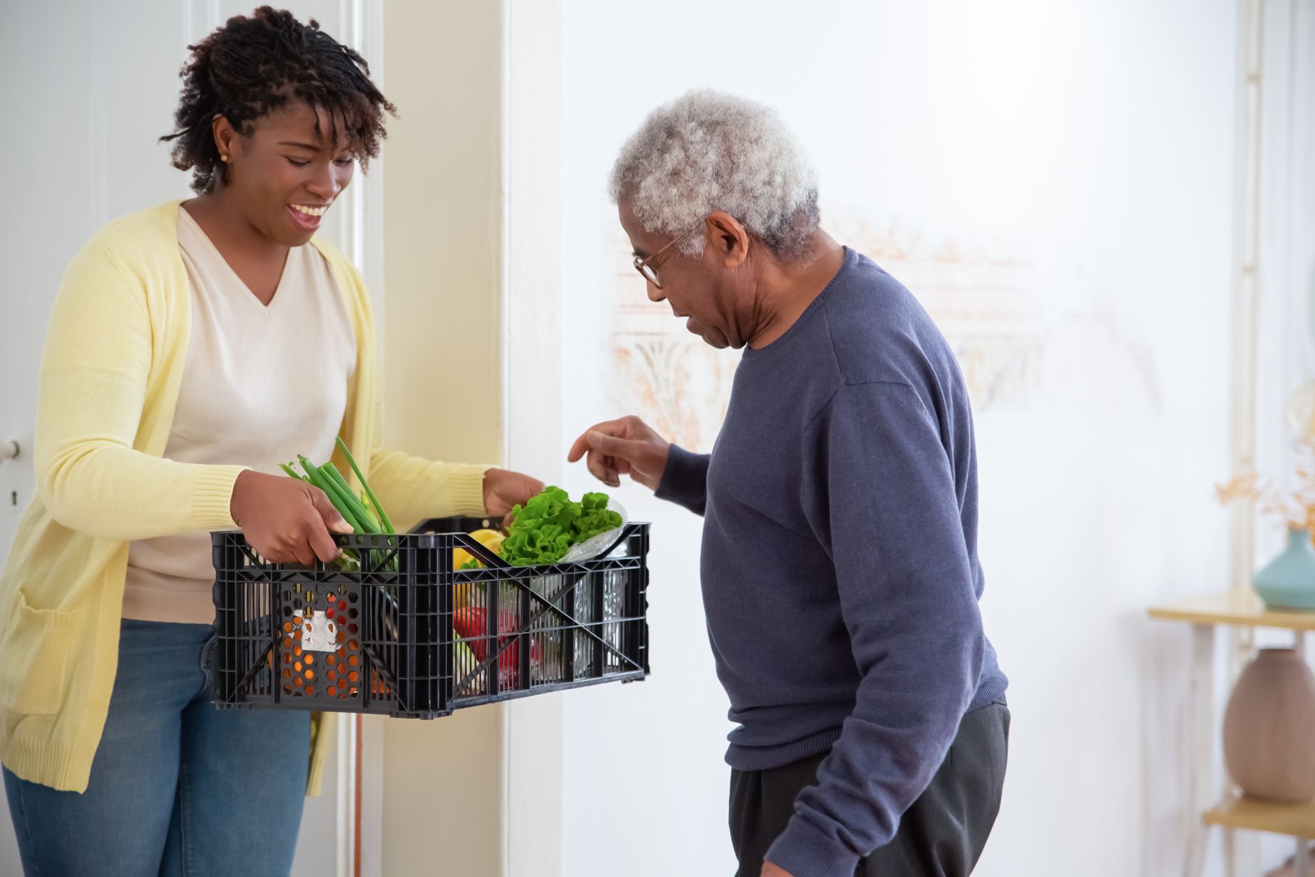 AI 助理 vs 人類助理:誰更懂你的需求? A young adult woman helps a senior man by carrying a crate of fresh vegetables indoors, symbolizing care and support.