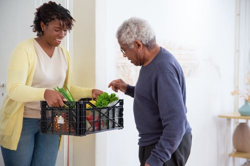 Free A young adult woman helps a senior man by carrying a crate of fresh vegetables indoors, symbolizing care and support. Stock Photo