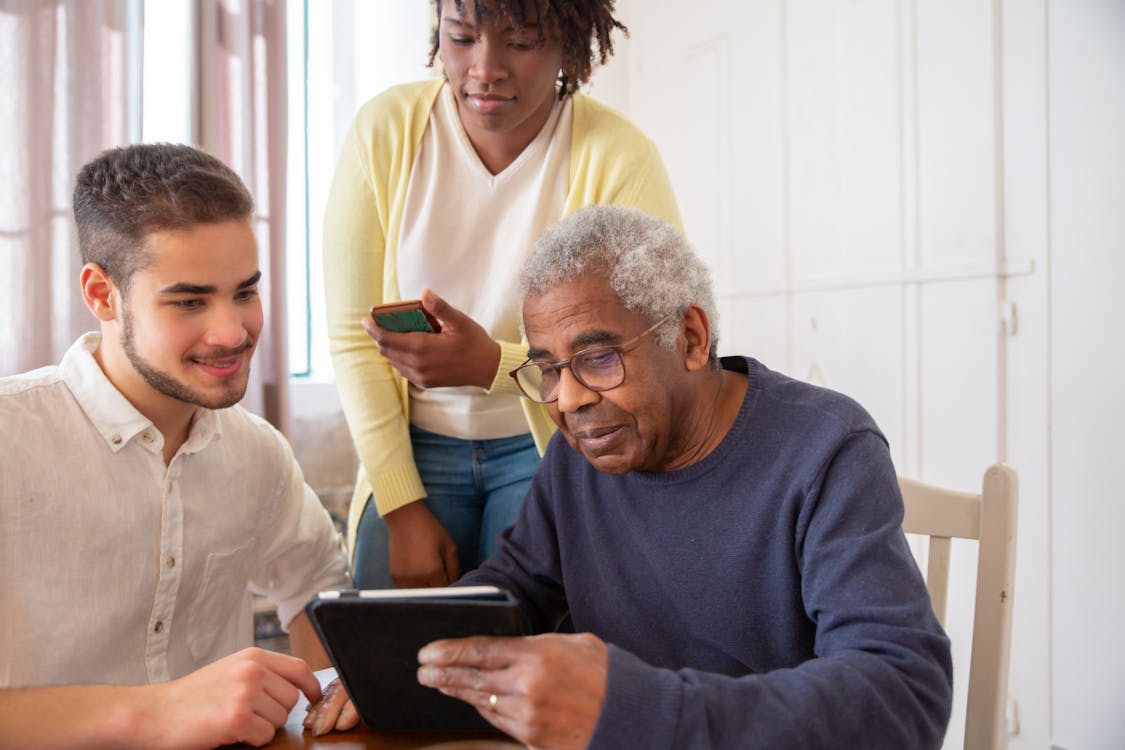 Free An Elderly Man in Blue Sweater Holding a Tablet while Talking to the People Beside Him Stock Photo