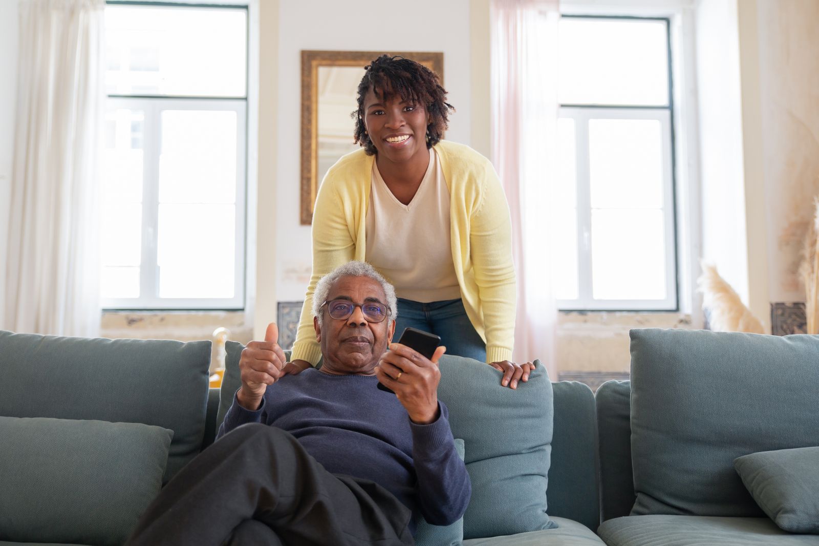 An older man on the sofa gives a thumbs-up while a family caregiver stands behind him in a bright living room.