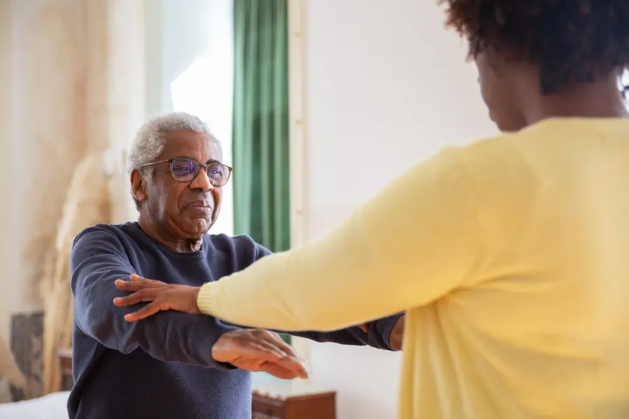 A carer gently supporting an older man as he stretches and moves around his home