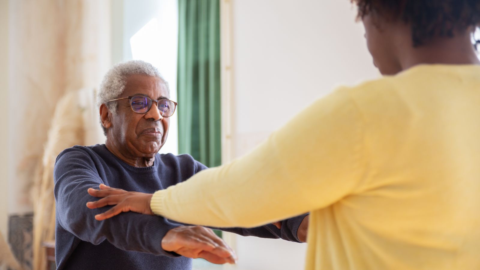 An elderly man with a caregiver assisting him in exercise at home.