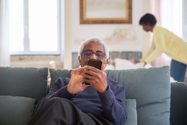 An Elderly Man Sitting On The Couch While Using His Mobile Phone