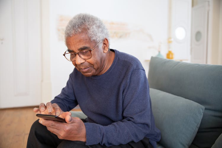 An Elderly Man Sitting On The Couch While Using His Mobile Phone