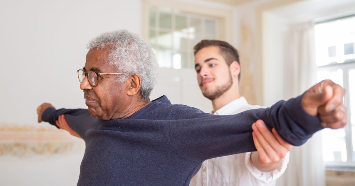 A senior man is assisted by a caregiver with a shoulder stretching exercise indoors
