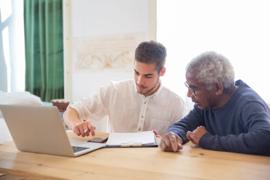 A care worker sitting at a kitchen table with an older man, going through paperwork together at home