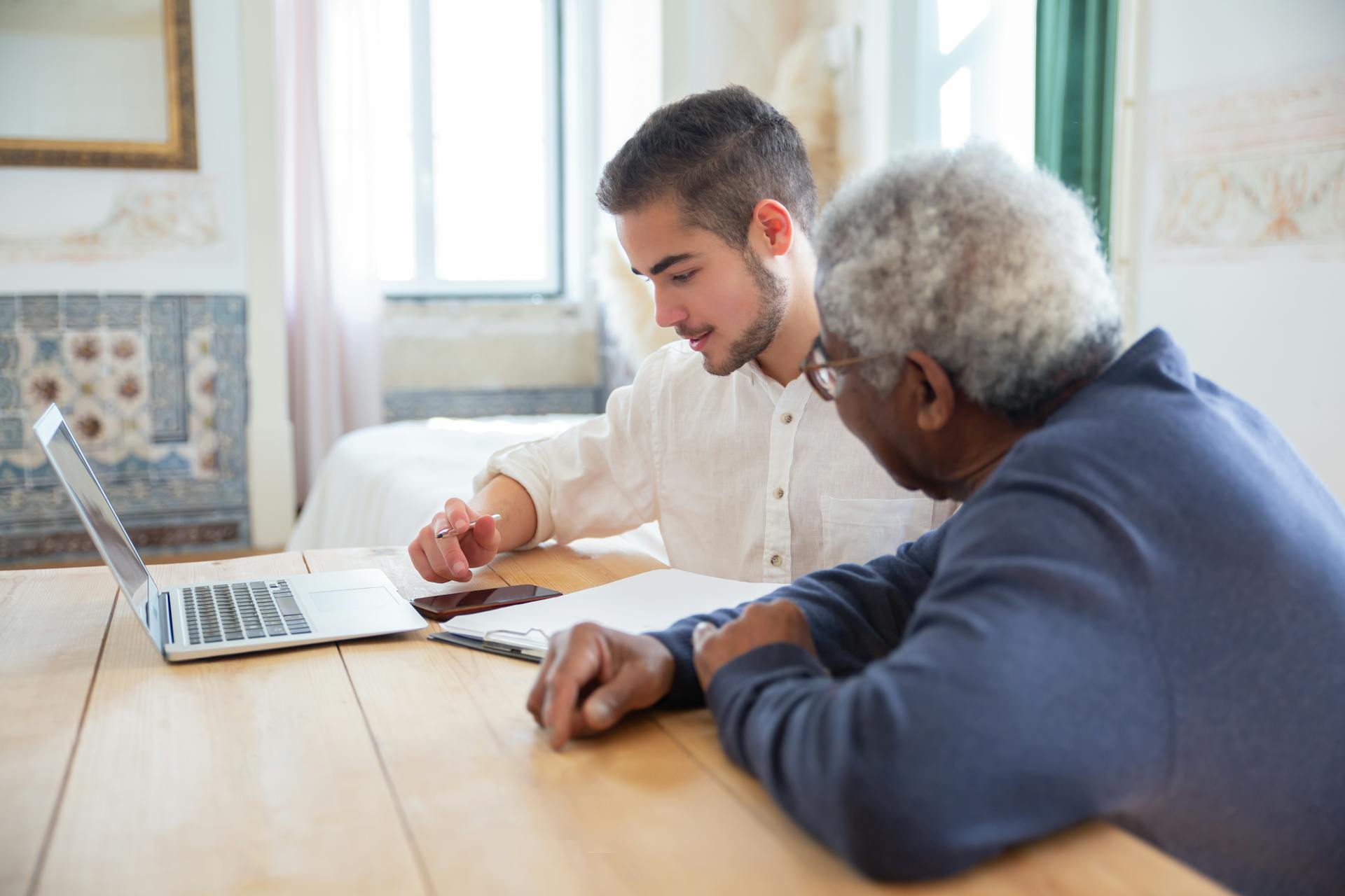 Young man assisting senior adult with laptop at home for social needs.