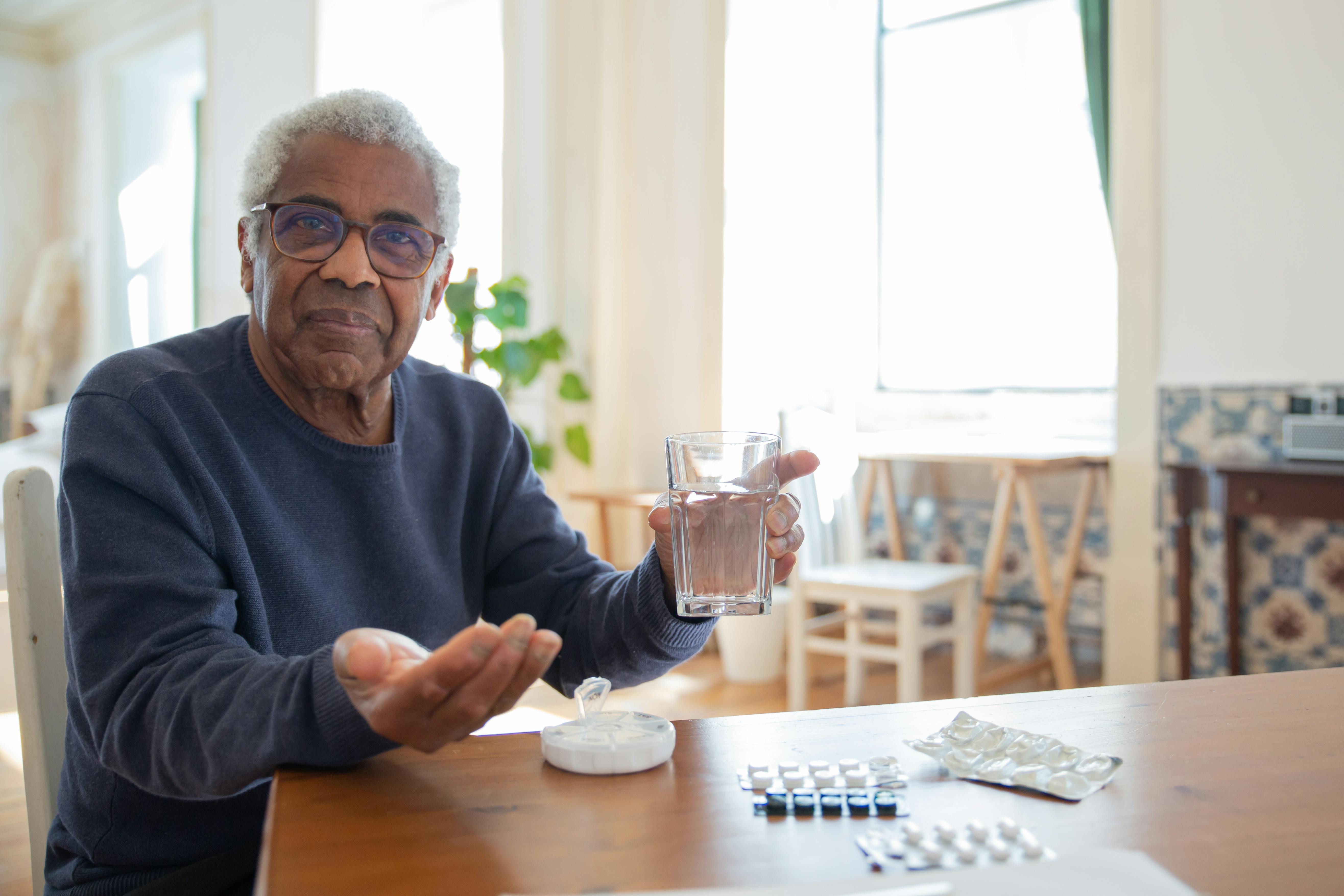 Free Elderly man sitting at a table at home, holding a glass of water and medication, emphasizing healthcare routine. Stock Photo