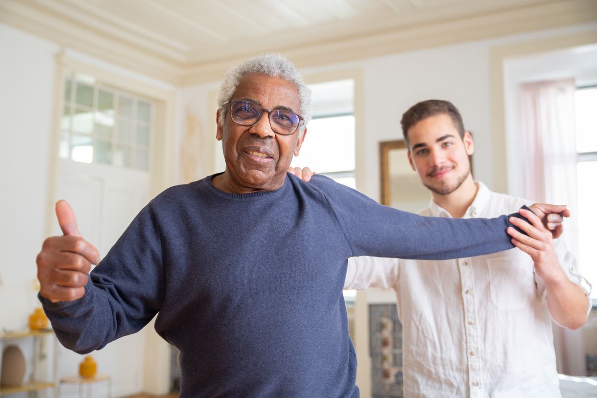 A smiling Black elder giving a thumbs up while a caregiver supports his arm