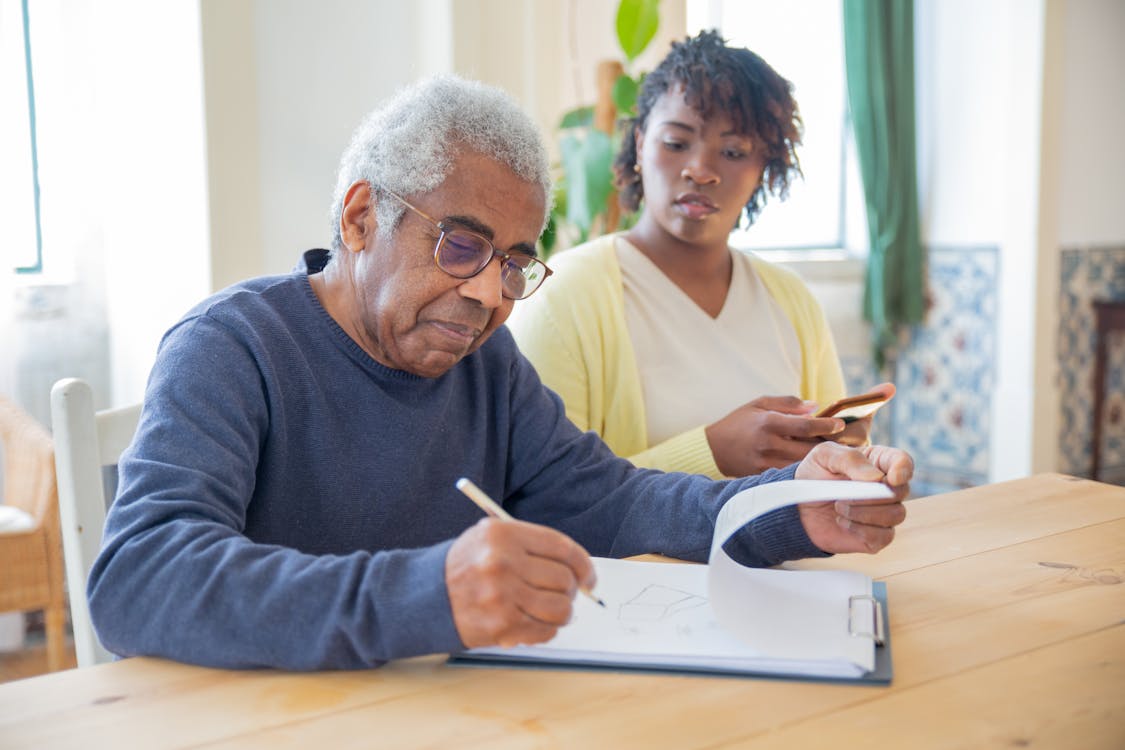 Free A senior man writing on papers with a caregiver nearby indoors in a supportive setting. Stock Photo