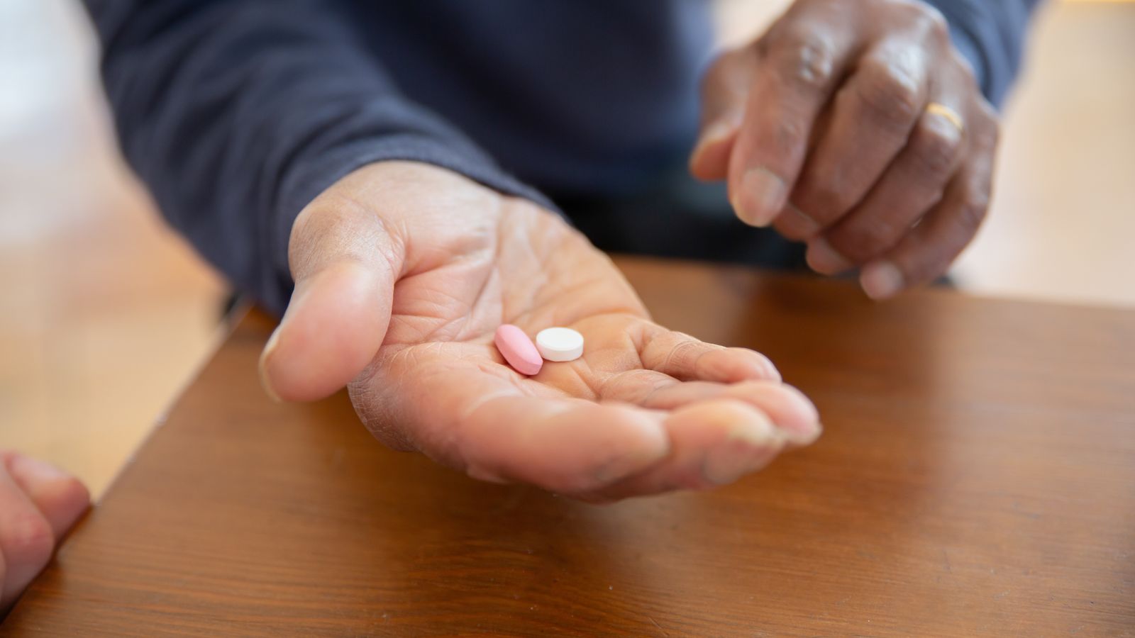 A close-up view of hands holding pink and white pills on a wooden table.