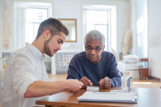 Young man assisting elderly man with medication in a bright living room setting.