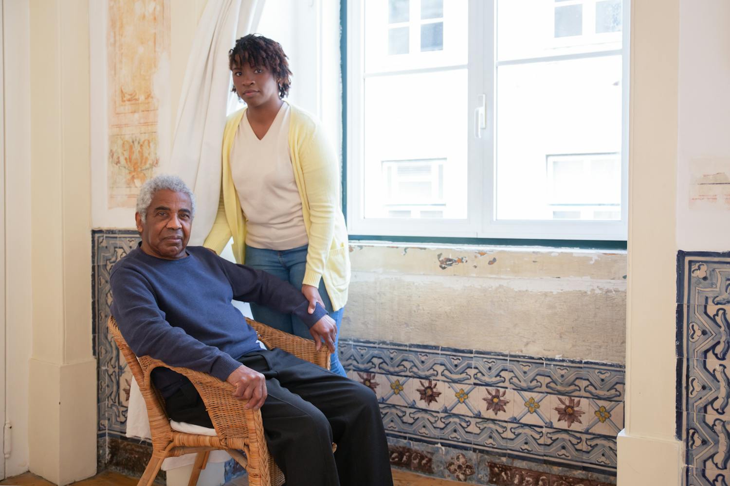 Elderly man seated with caregiver standing beside him at home