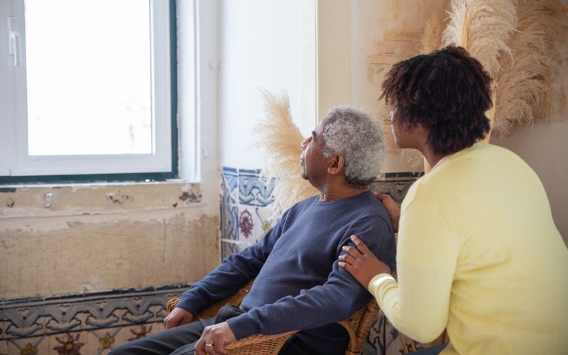 A caregiver sitting close beside an older man, both looking toward the window
