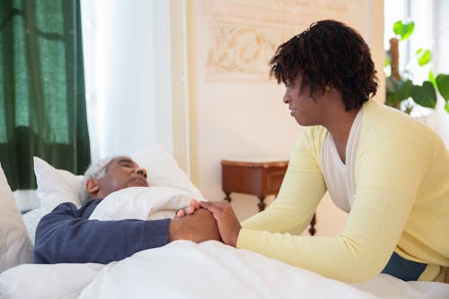 Woman in Yellow Cardigan Sitting beside a Man in Blue Long Sleeve Shirt Lying on Bed