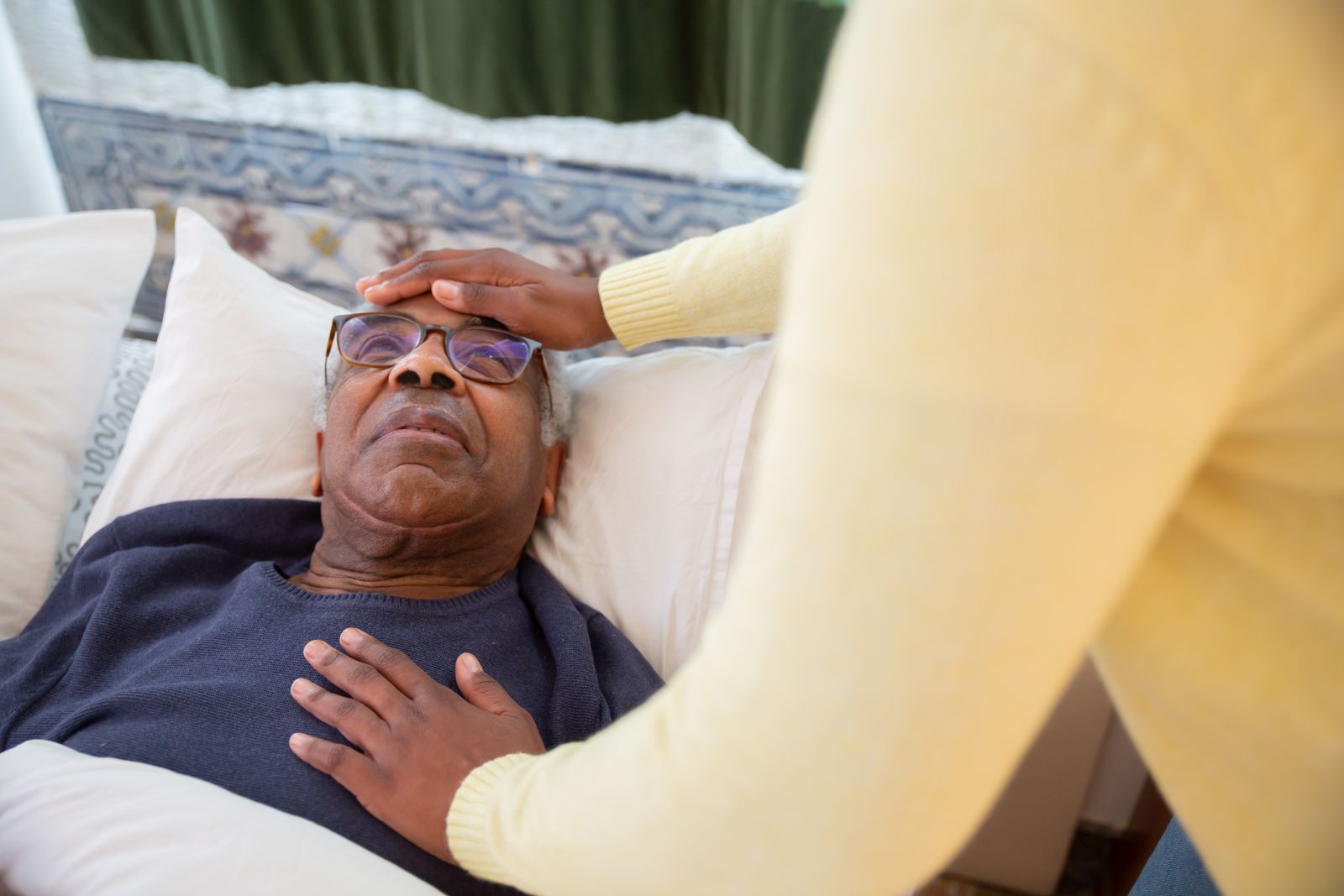 A younger caregiver checks on an older man resting in bed at home, the kind of presence that still needs a person.