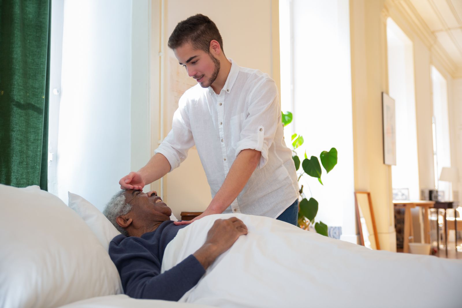 A younger caregiver checks on an older man resting in bed at home, the kind of presence families pay for by the hour.