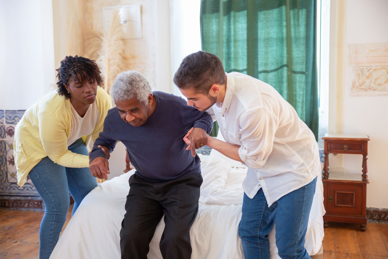 Two people help an older man stand up from the edge of a bed, the kind of transfer that still needs human hands.