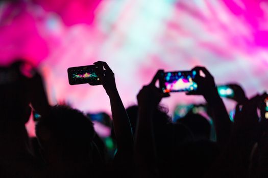 Silhouettes of people recording a vibrant concert with mobile phones, capturing the colorful stage lights.