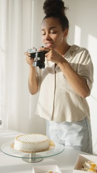 A young woman captures a photo of a cake on a cakestand in a well-lit room.