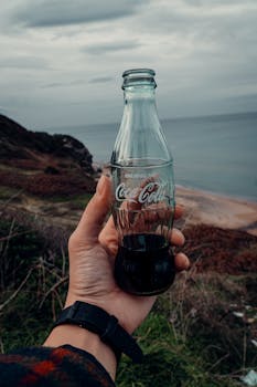 A hand holding a Coca-Cola glass bottle with a scenic outdoor background.