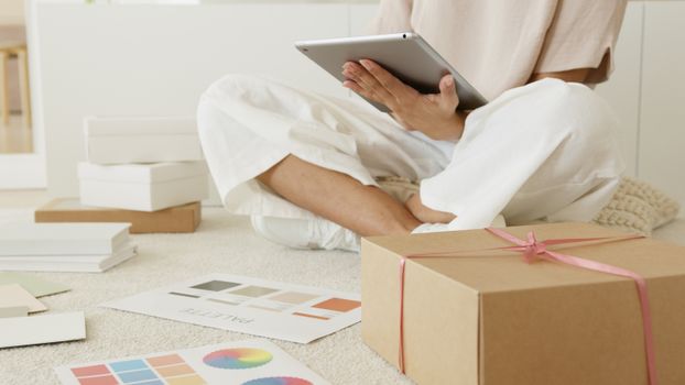 A person sitting with a tablet, surrounded by gift boxes and color palettes, in a cozy indoor setting.