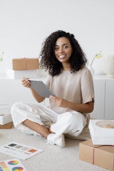 African American woman sitting with a tablet, surrounded by parcels, with a cheerful expression.