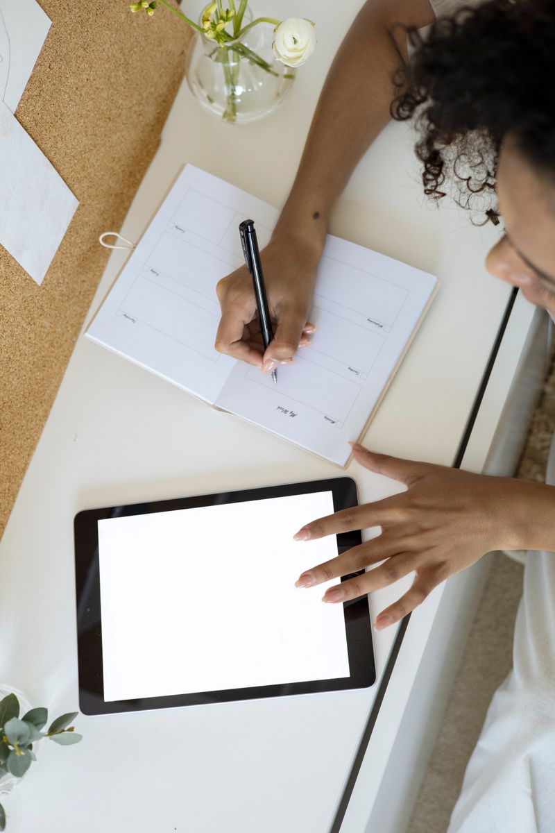 Woman uses a tablet and notebook for planning at a white desk