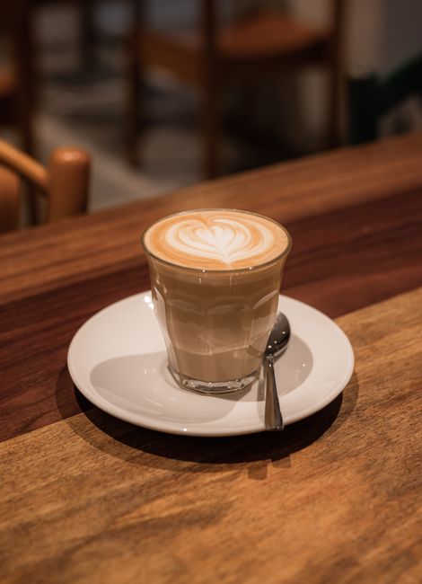 A cappuccino with heart latte art in a clear glass on a saucer atop a wooden table.