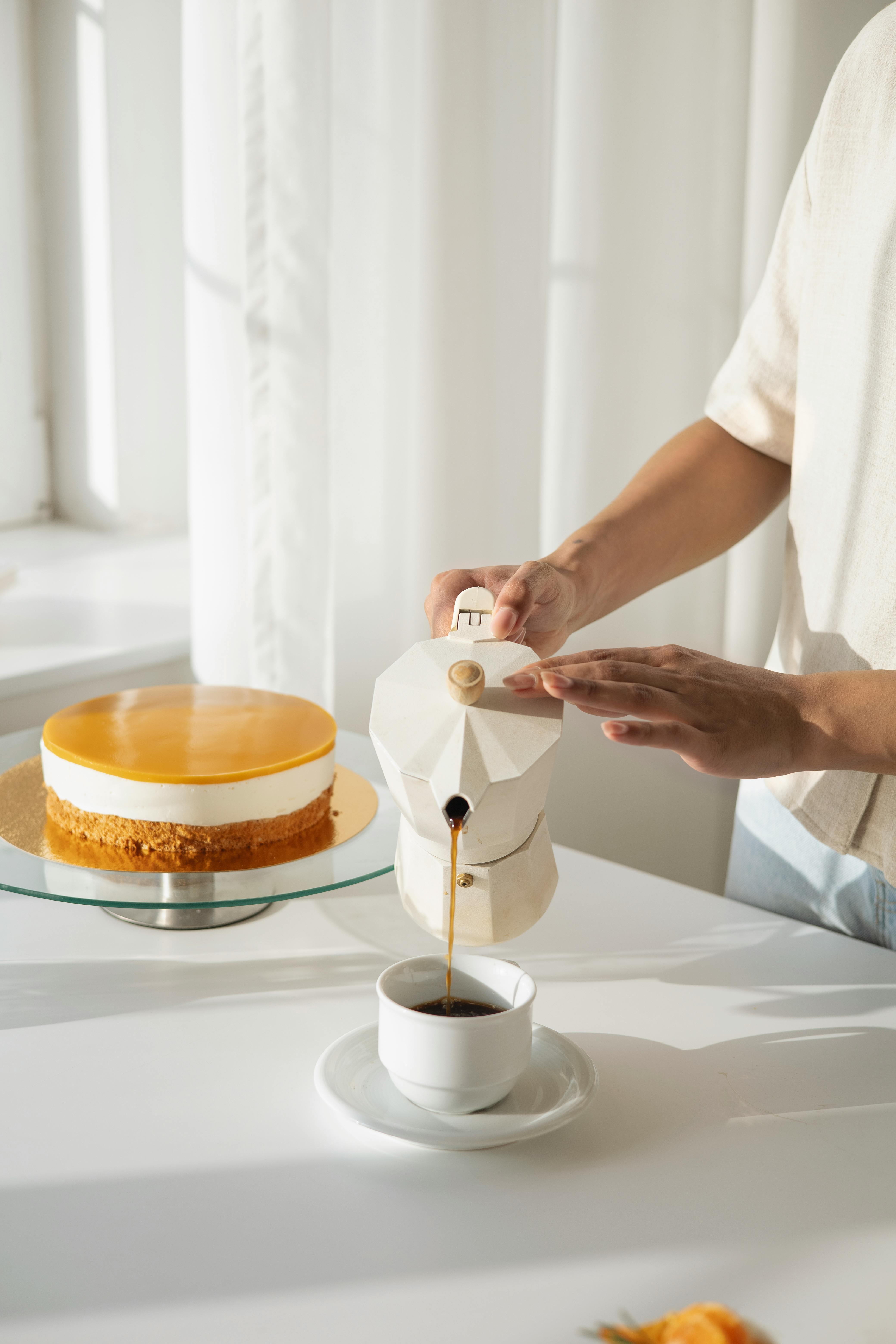 A person pours coffee from a ceramic kettle with a cake on the table, in a bright kitchen setting.