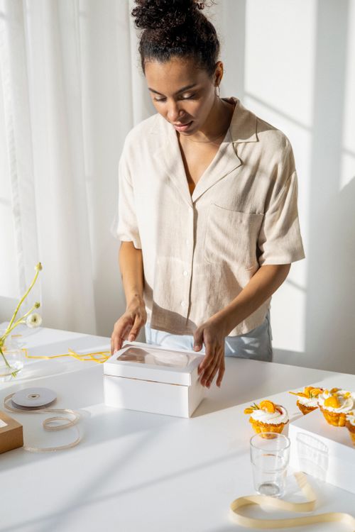 Free Black woman preparing a gift box indoors with cupcakes and ribbon, radiating a sunny mood. Stock Photo