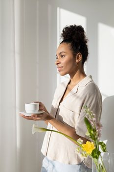 Smiling woman with curly hair holding a cup and saucer by a sunlit window indoors.