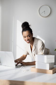 A smiling businesswoman using a laptop indoors with natural light and boxes on the table.